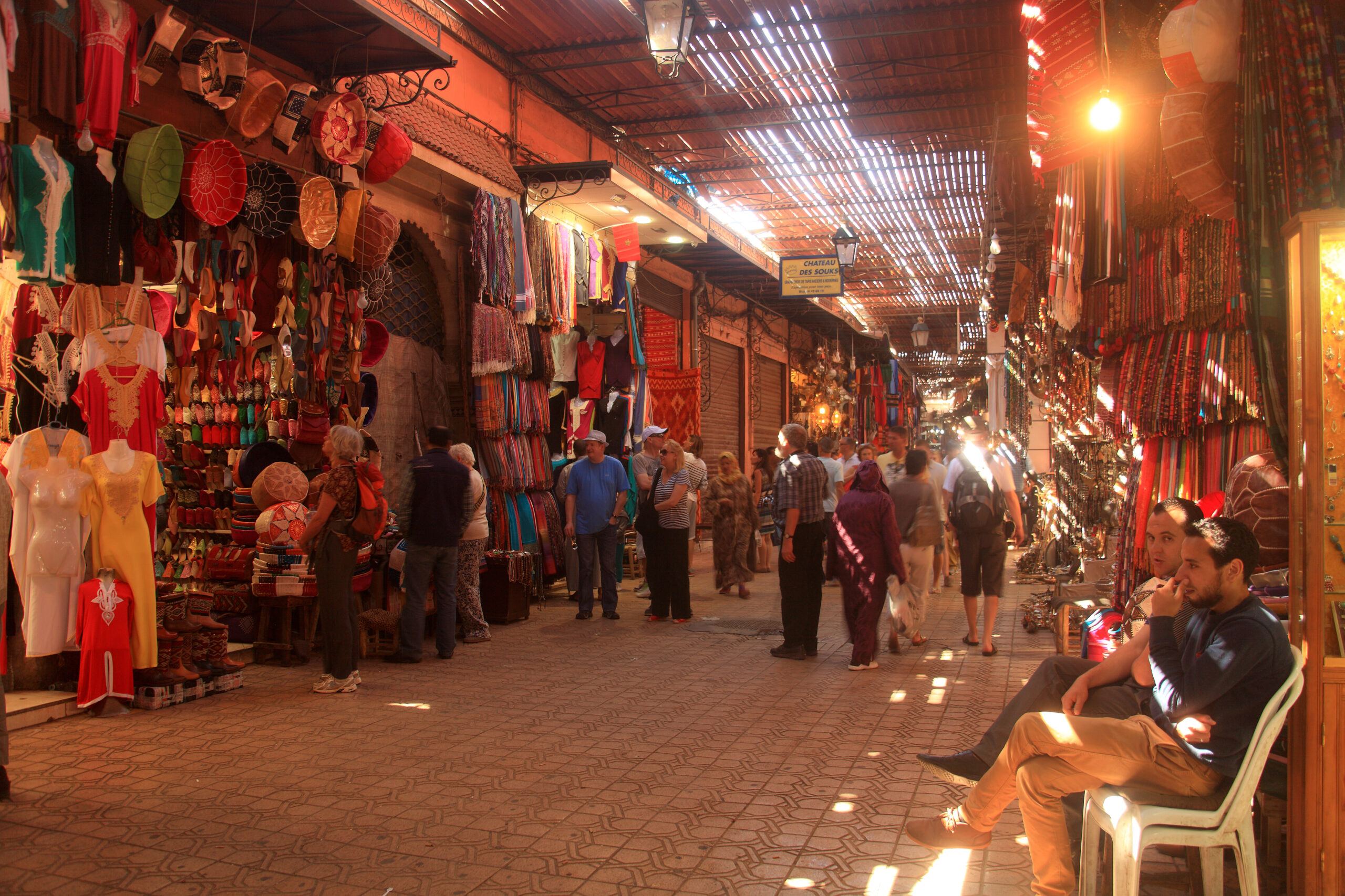 Visitors walking through a covered souk alley in the Marrakech medina, with shaded stalls and level paving